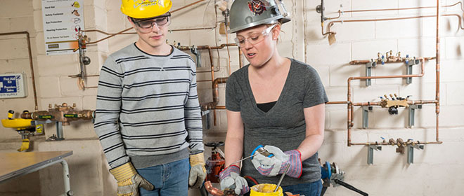 a female plumber journeyperson training a male apprentice on how to solder a copper pipe.
