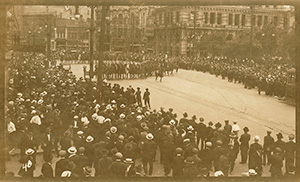 a street in 1919 in Winnipeg. A large crowd of people stand on both sides of the street. Two rows of police offers on horses march down the middle of the street.