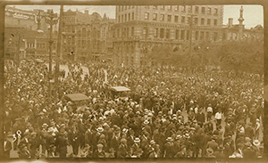 A large crowd of people standing in the street in 1919 in Winnipeg. There are two automobiles in the midst of the crowd. Buildings line the street in the background.