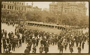 A large crowd of people pushing a trolly car over onto its side.