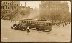 The large crowd is gone, with come people in the background. The trolly car that had been pushed onto its side is now upright. Smoke billows from the trolly. An automobile and three men stand next to it. 