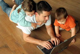 man lying on the floor with a child beside him and on his back, all are looking at a computer