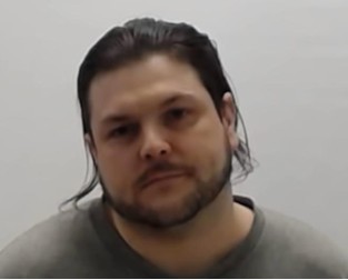 White male, dark brown long hair chin length, short beard, grey shirt, standing against a white wall